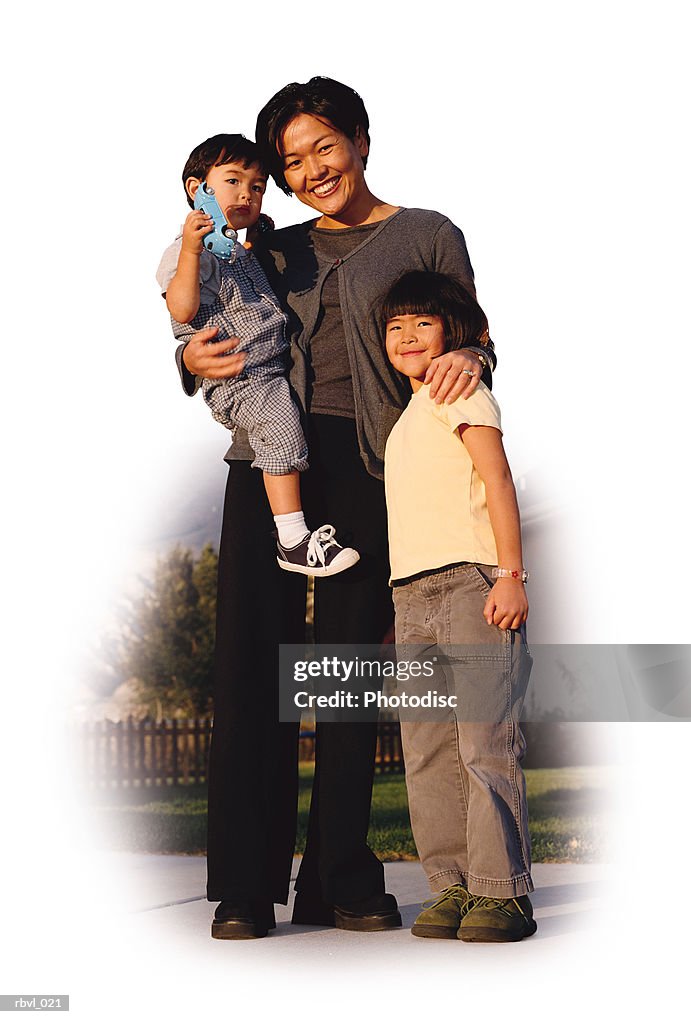 A young asian mother wearing dark pants and blouse is holding her toddler son and has her arm around her little girl as they smile towards the camera