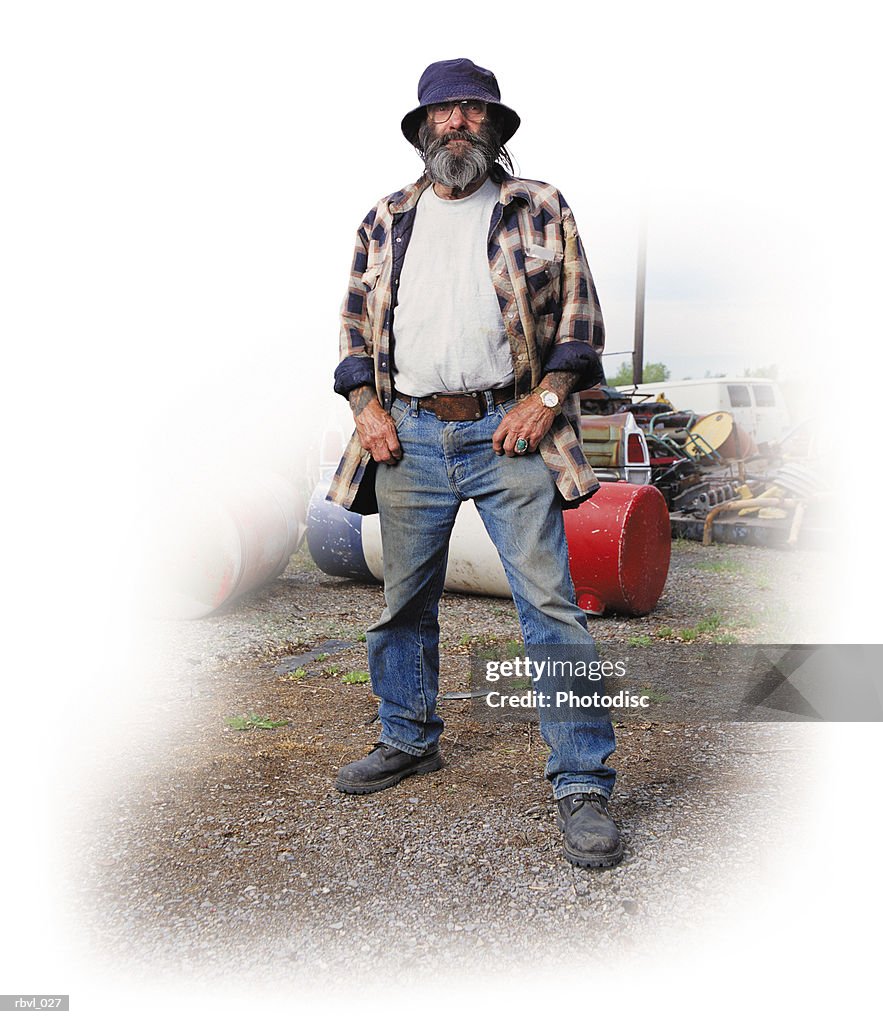 A casually dressed older caucasian man with long facial hair wearing dirty jeans and a floppy hat is standing in a yard full of junk