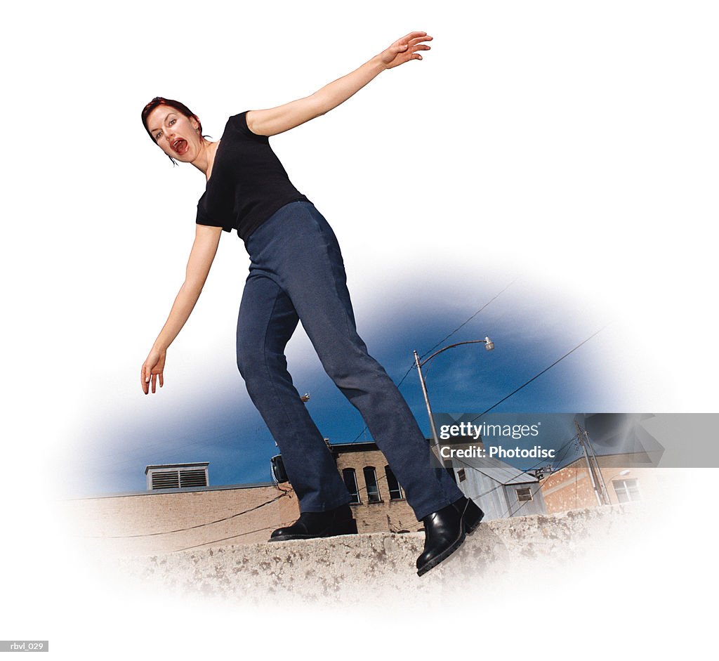 A young red haired caucasian woman in black boots wearing jeans and a black shirt is balancing along the top of a wall that is in front of some old buildings