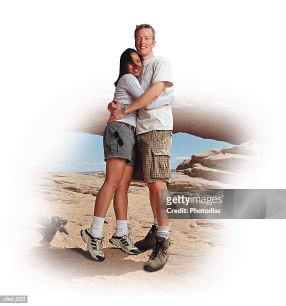 a young caucasian man in checkered shorts and a white t-shirt is hugging a young caucasian woman in gray shorts and a long sleeved white shirt in front of a natural arch in the southern utah desert - white shirt ストックフォトと画像