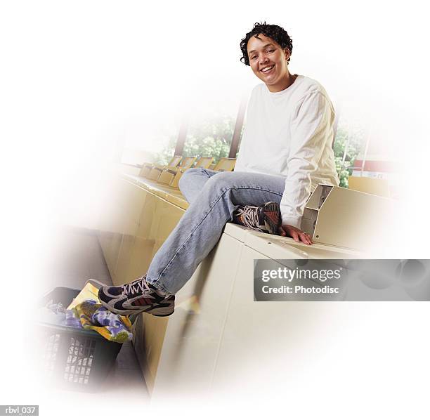 a young african american woman in blue jeans and a white long sleeved shirt is sitting on top of a washing machine inside a laundromat - inside of ストックフォトと画像