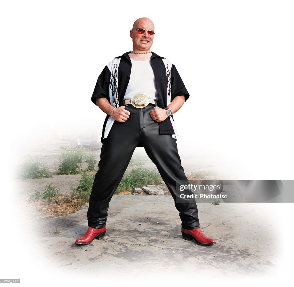 A punk bald caucasian cowboy in leather pants and red snakeskin boots with red tinted sunglasses is standing in front of an abandoned factory with a sneer on his face