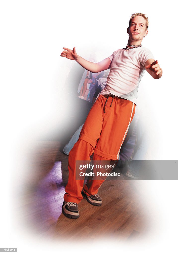 A young caucasian man with a goatee and spiked blond hair wearing orange pants and a white t-shirt is dancing in a nightclub