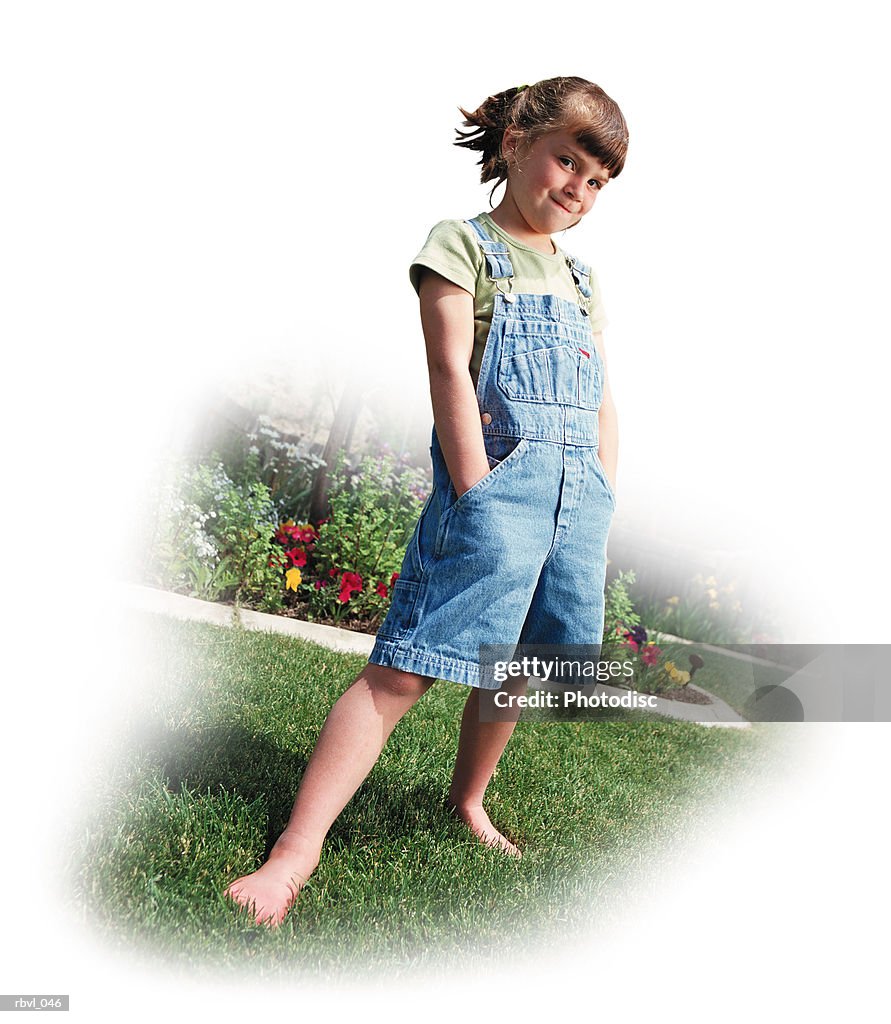 A little caucasian girl with a ponytail is wearing short overalls with her hands in her pockets as she stands on a lawn bordered by flowers