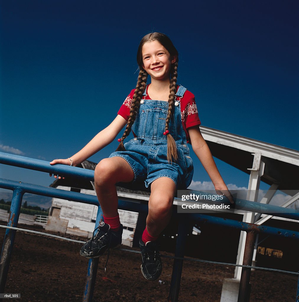 A young girl in jean overalls and red shirt with long braids sits on a fence smiling at the camera