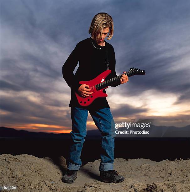 a male caucasian youth with long blond hair wearing a black shirt and blue jeans is standing outside playing an electric guitar - modern rock stock pictures, royalty-free photos & images