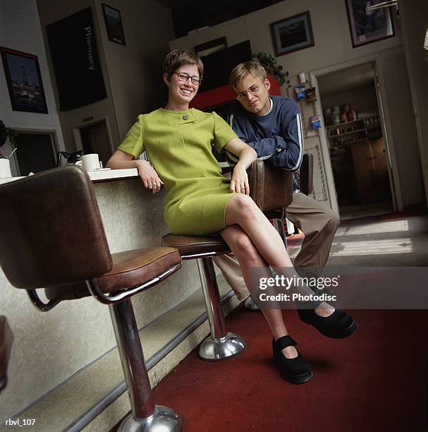 a young caucasian man in tan pants and a long sleeve blue shirt and a young caucasian woman in a short green dress are sitting on barstools in a cafe looking into the camera - dress short sleeve stockfoto's en -beelden