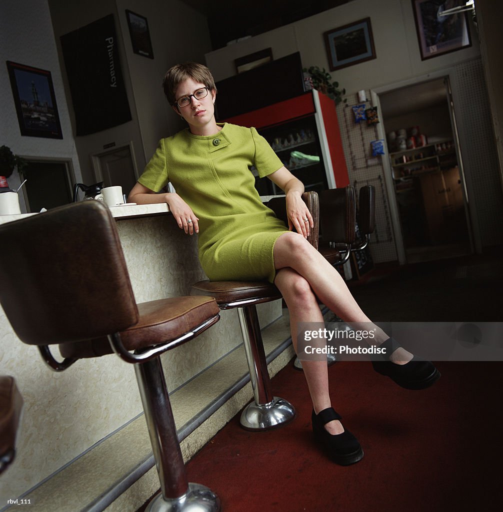 A young caucasian woman with a short trendy green dress and glasses is sitting at the bar of a diner looking seriously at the camera