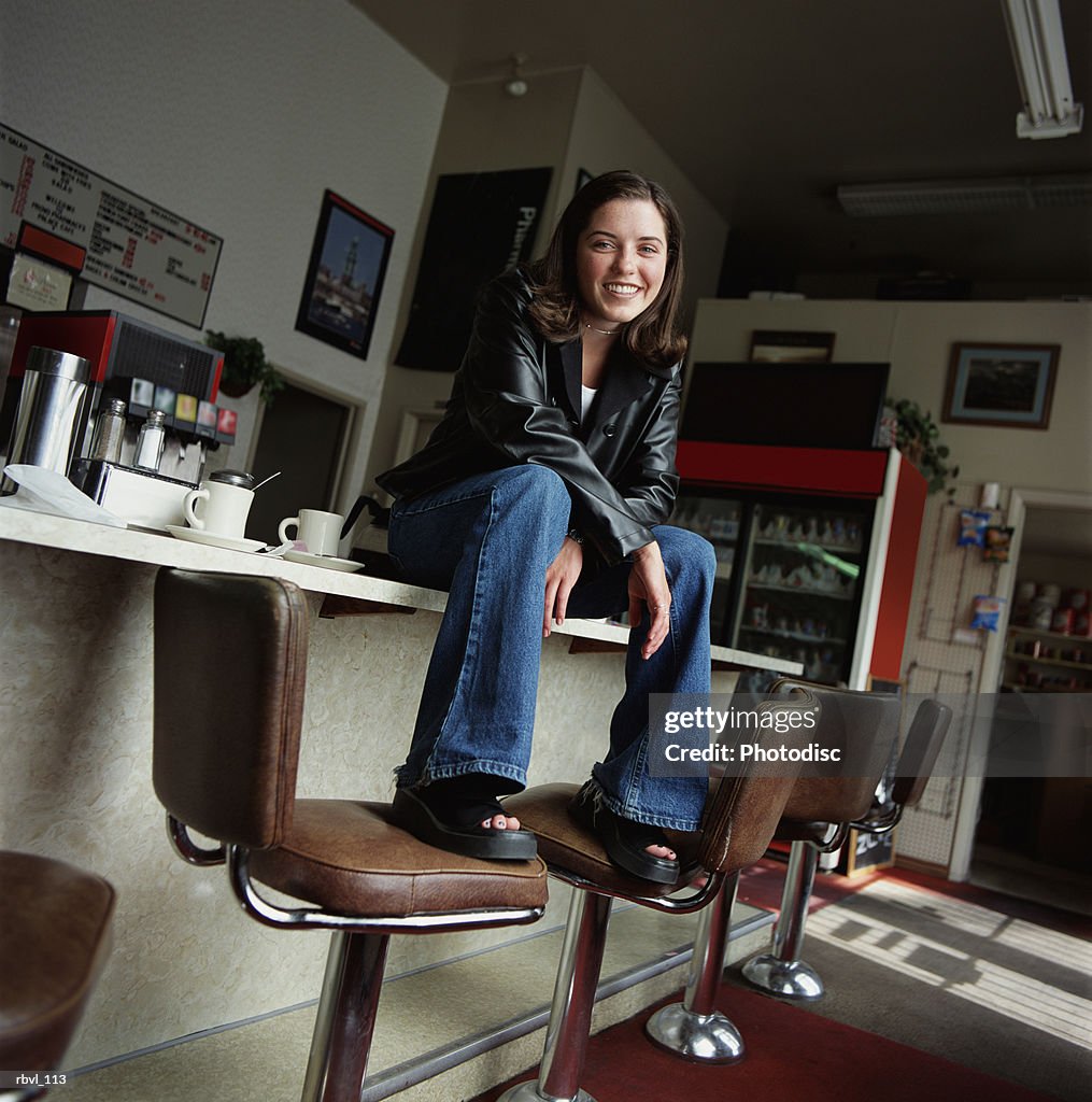 A young hispanic woman in blue jeans and black leather jacket is sittng on the counter of a diner looking into the camera