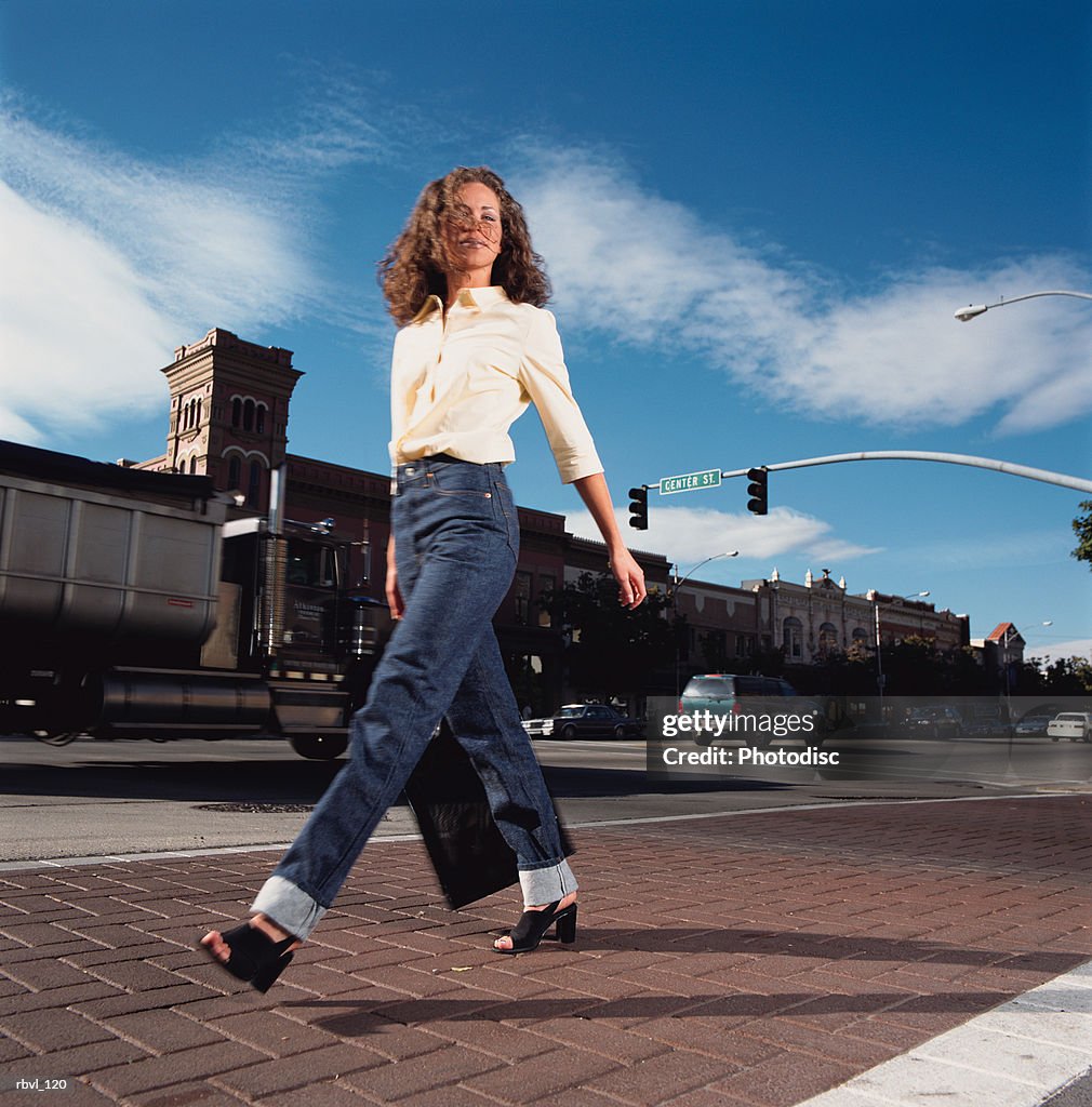 A young hispanic or caucasian woman with curly brown hair in a yellow shirt and blue jeans is walking along a city street