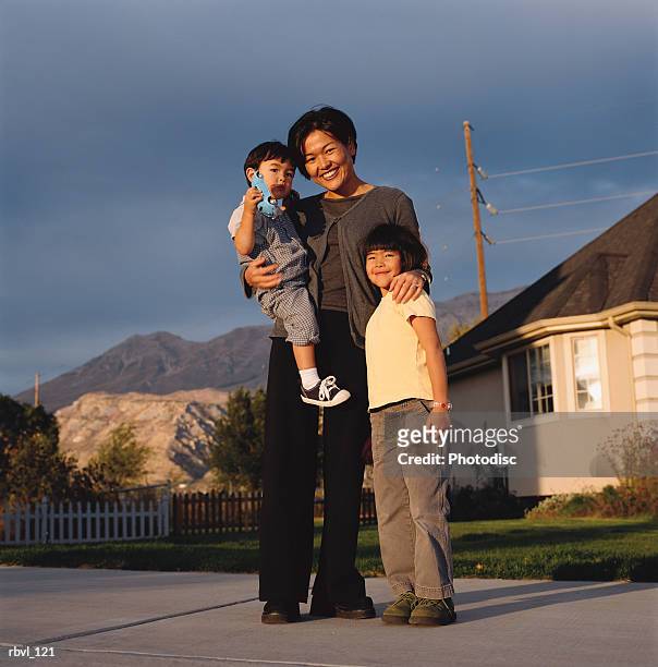 a young asian mother wearing dark pants and blouse is holding her toddler son and has her arm around her little girl as they smile towards the camera - smile imagens e fotografias de stock