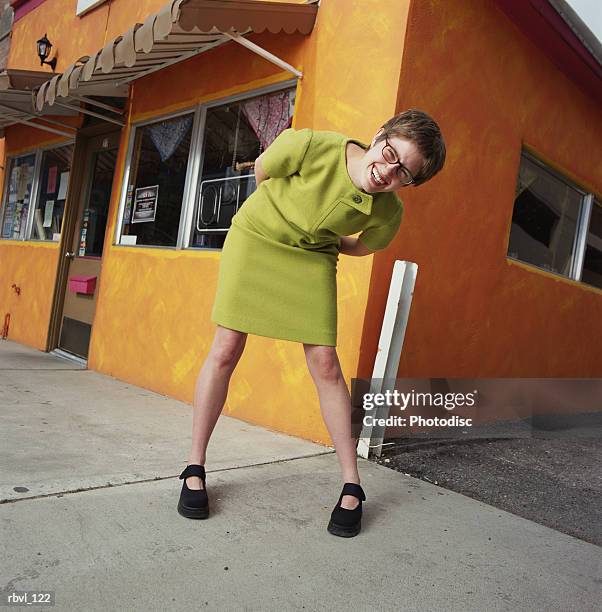 a laughing young caucasian woman in a short trendy green dress is bending down to look into the camera with an orange building in the background - look down stock-fotos und bilder