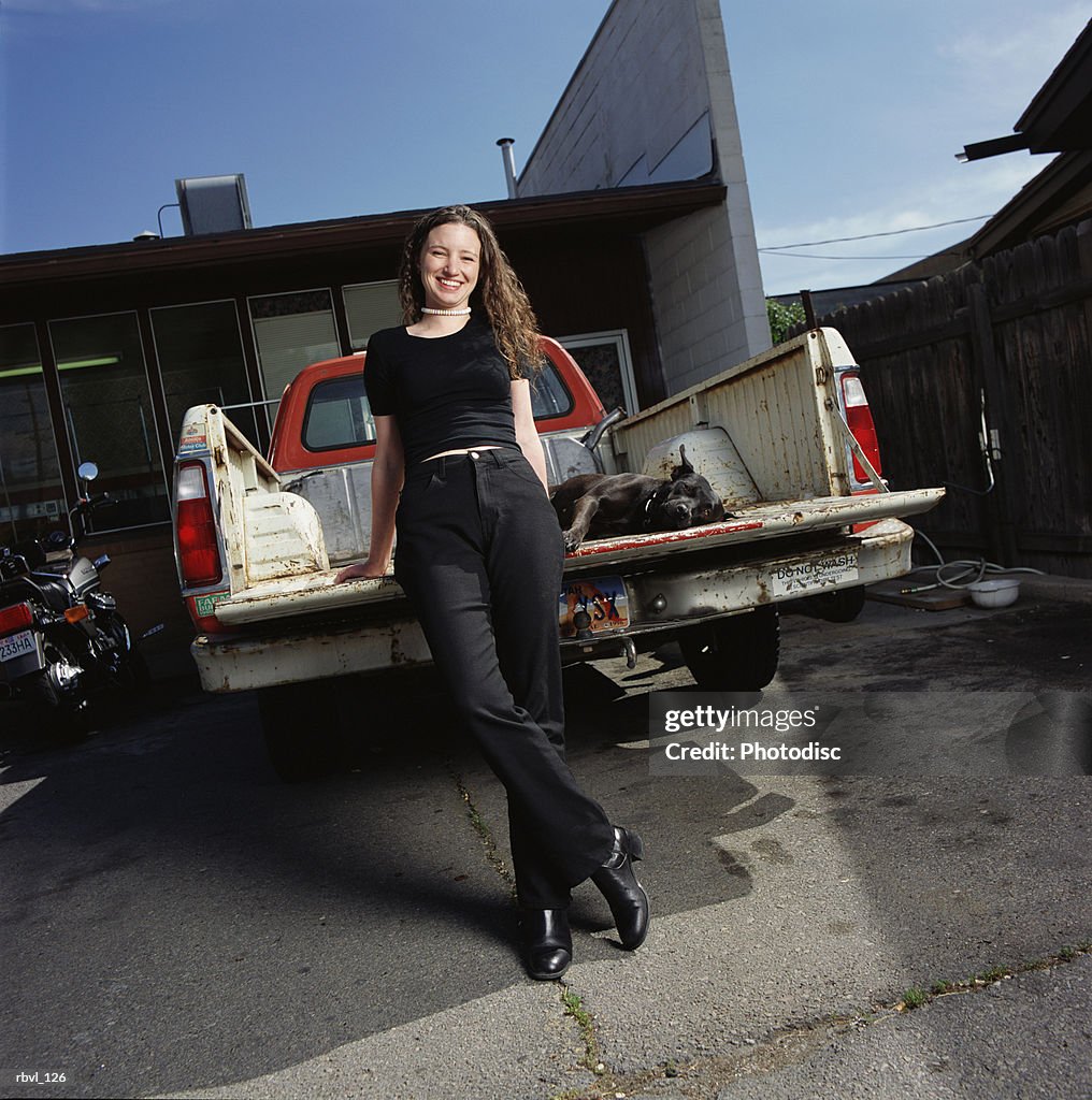 A young caucasian woman in black pants and shirt is leaning against the tailgate of a pickup truck with her black dog lying in the truck bed