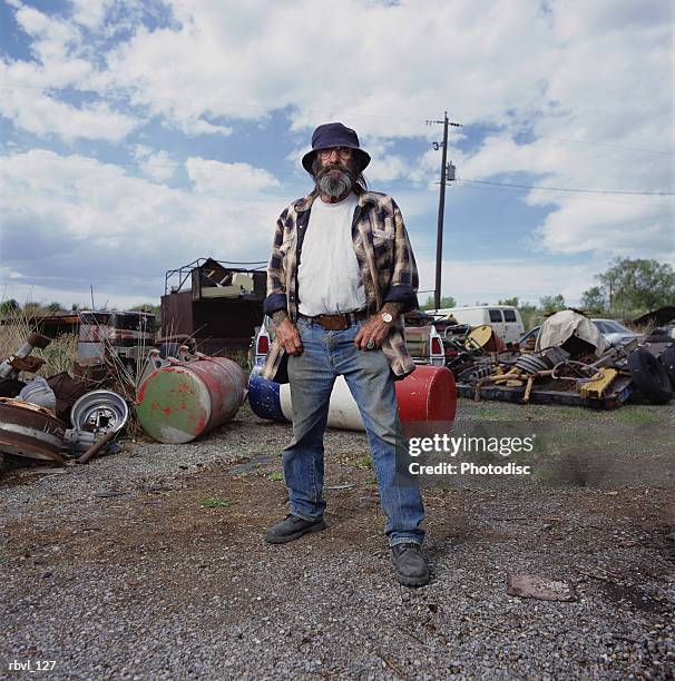 a casually dressed older caucasian man with long facial hair wearing dirty jeans and a floppy hat is standing in a yard full of junk - junk ストックフォトと画像