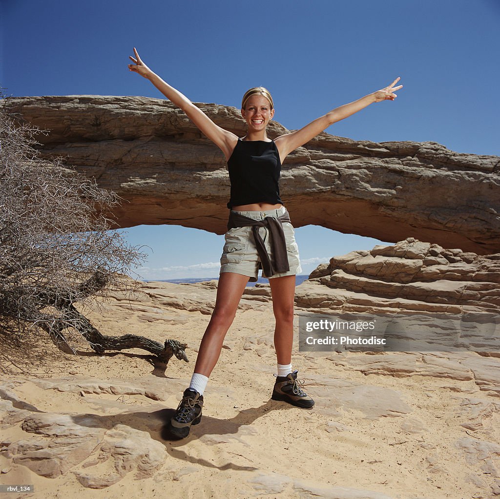 A young blond caucasian woman is wearing shorts and a tank top in front of a natural arch with her arms extended in a peace gesture