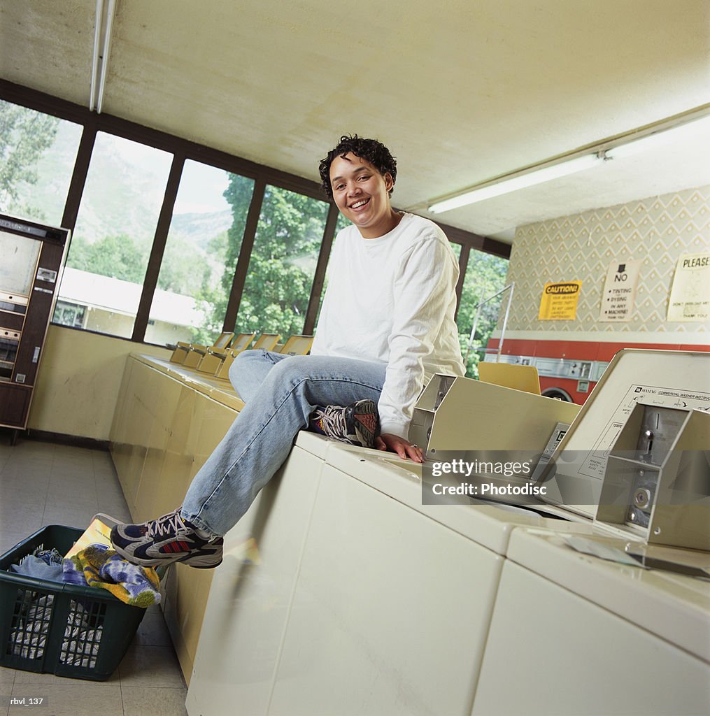 A young african american woman in blue jeans and a white long sleeved shirt is sitting on top of a washing machine inside a laundromat