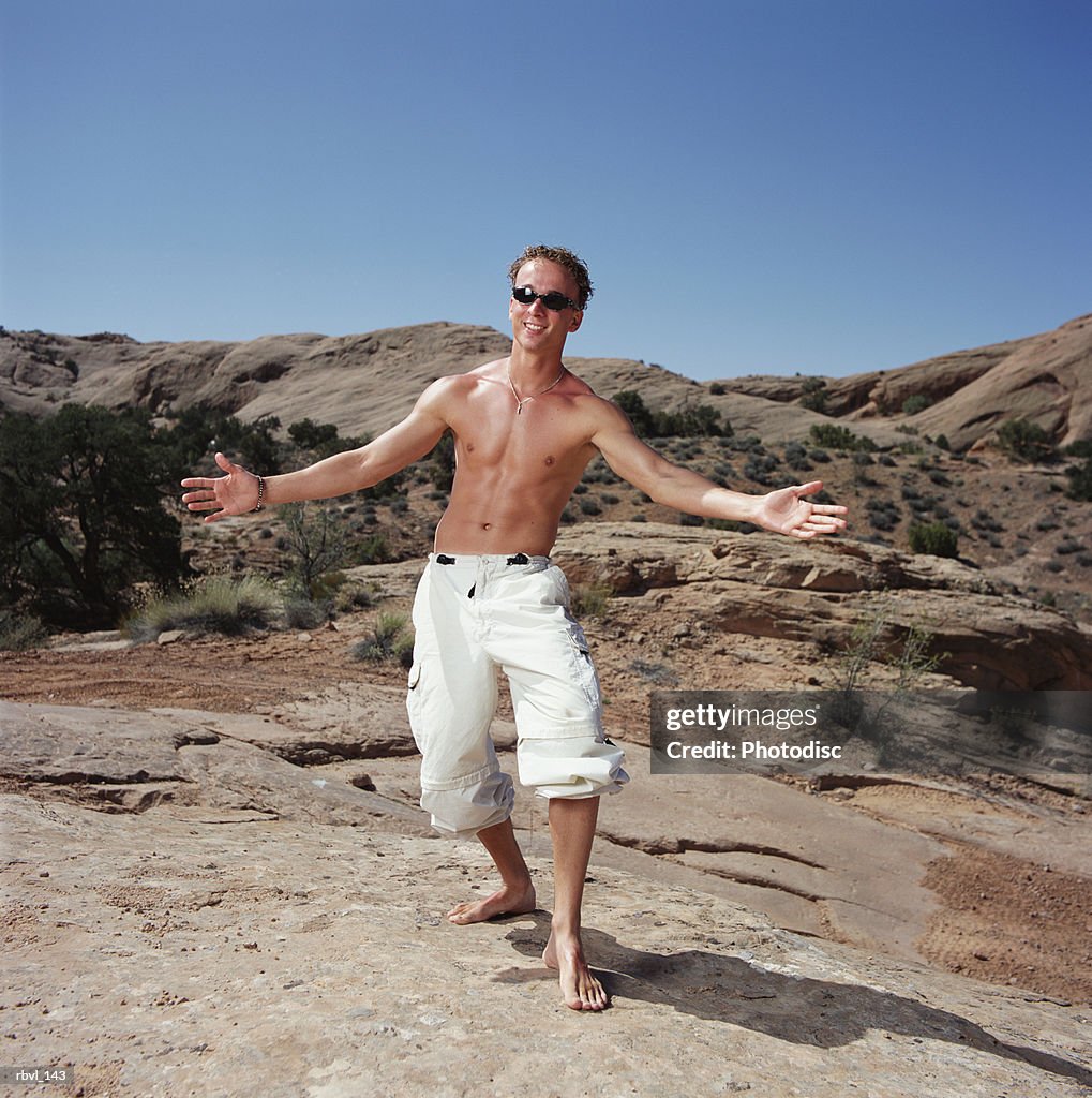 A tan caucasian young man in sunglasses is wearing baggy white pants and no shirt as he stands gesturing with outstretched arms toward the camera