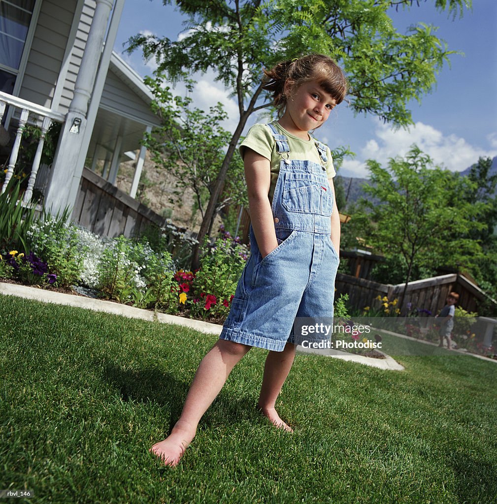 Caucasian girl with ponytail wears short overalls hands in pocket stands on lawn bordered by flowers