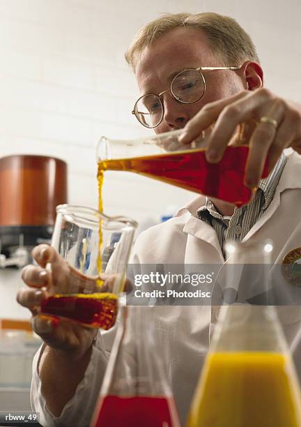 a caucasian chemist in a laboratory pours colorful liquids from one beaker to another - aother stock pictures, royalty-free photos & images