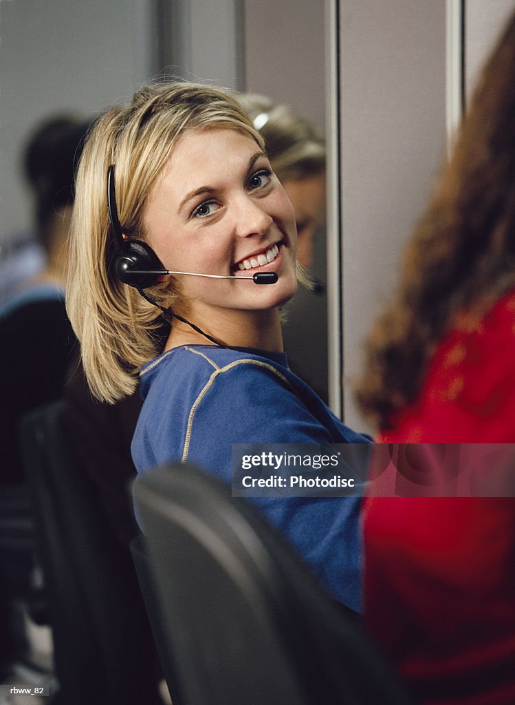 A blonde female telemarketer wearing a headset sits at her cubicle and smiles