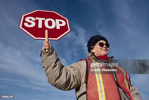 a female caucasian wearing sunglasses and a wool cap holds a stop sign at a crosswalk - 交通誘導員 ストックフォトと画像