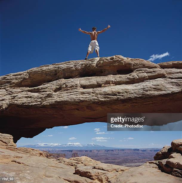 a young bare chested man stands triumphantly on the top of a natural arch in the south utah desert - valley type stock pictures, royalty-free photos & images