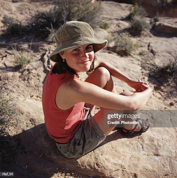 a young woman i n grey shorts a red shirt and tan hat is sitting on a rock in the south utah desert - is stock pictures, royalty-free photos & images