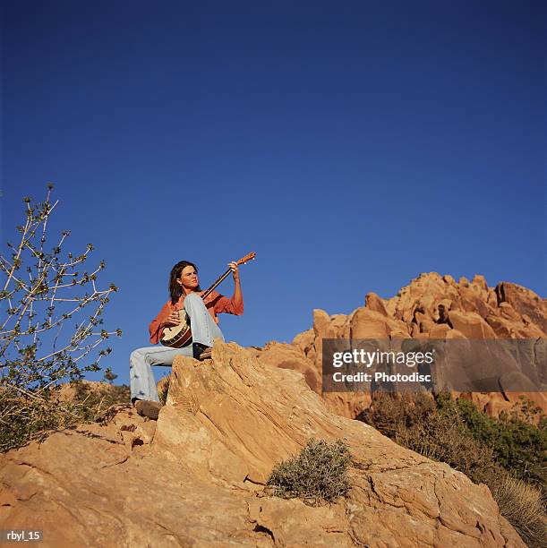 a young woman in blue jeans and a red long sleeved shirt is sittingn on the crest of a boulder playing a banjo with the blue sky and other rock croppings in the background - boulder rock stock-fotos und bilder