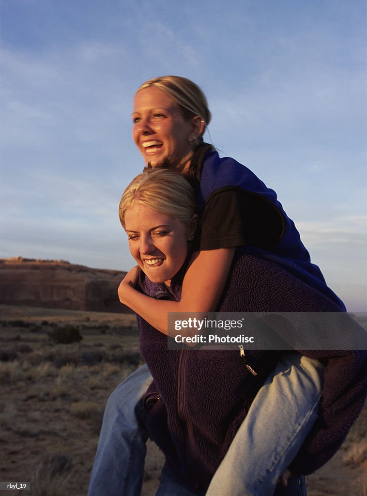 A young woman in blue jeans and a purple fleece is giving her friend who is wearing blue jeand and a blue fleece vest a piggyback ride with the southern utah desert in the background