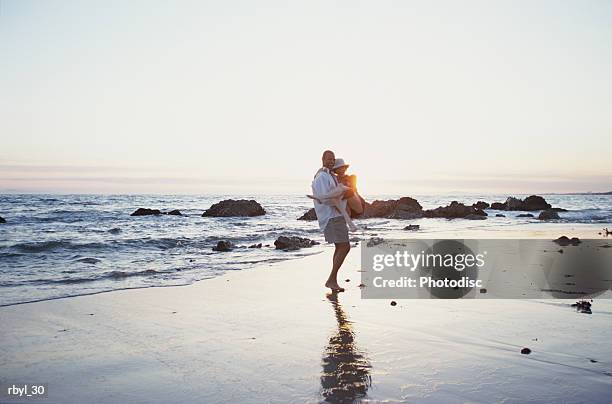 a youing man is carrying a young woman in his arms on a rocky beach at sunset with the ocean behind them - is stock pictures, royalty-free photos & images