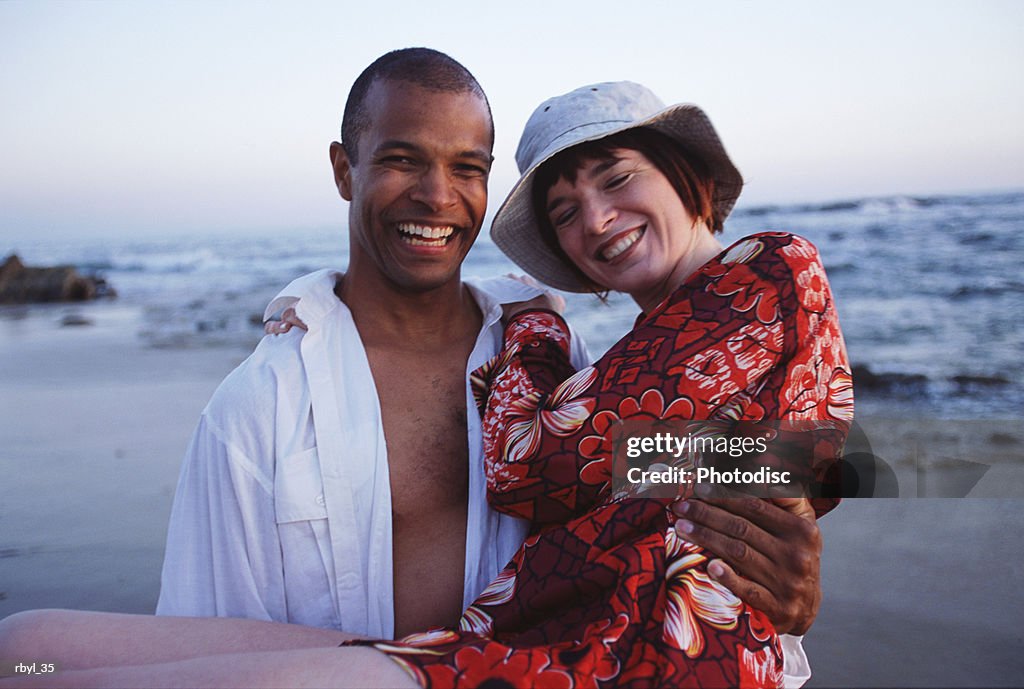 A young man is carrying a young woman in his arms with the beach and shoreline behind them