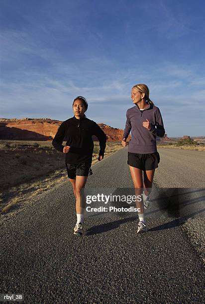 two young women in shorts and long sleeve shirts are running along a road in the south utah desert - long - fotografias e filmes do acervo