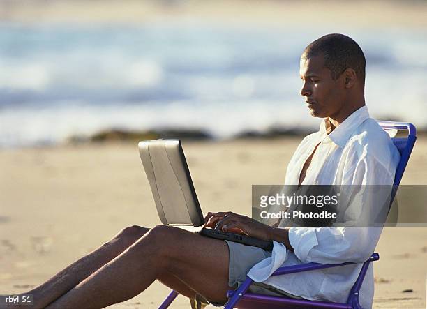 a young man in tan shorts and a white oxford shirt is reclining in a beach chair typing in to a laptop computer - tan tan stock pictures, royalty-free photos & images
