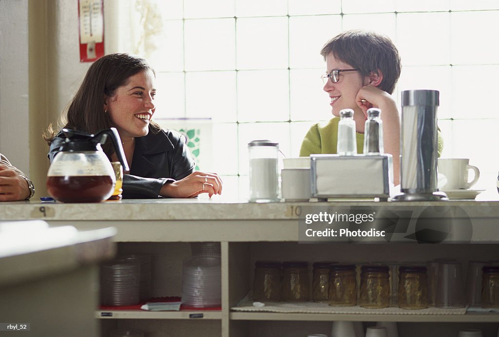 Two young women are sitting at the counter of a restauraunt or cafe talking to eachother