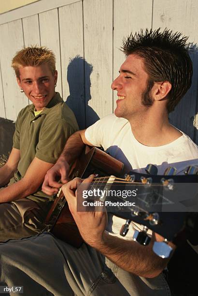 two teenage boys are sitting on a city sidewalk playing the guitar and talking to eachother - rock moderno foto e immagini stock