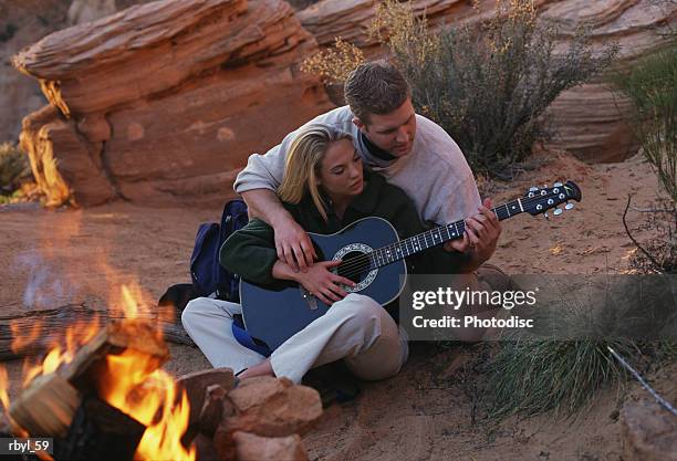 a young couple is sitting closely together beside a campfire where the man is showing the woman how to play the guitar with the desert  in the background - how fotografías e imágenes de stock