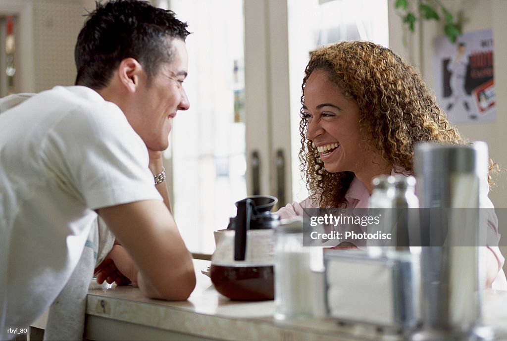A young man working the counter of a restuarant or cafe is laughing and tlaking with a young woman with curly brown hair