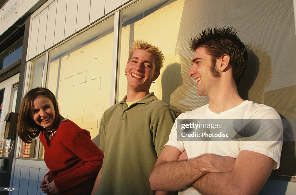 A group of two young men and a young woman are standing outside a city storefront laughing and talking to eachother