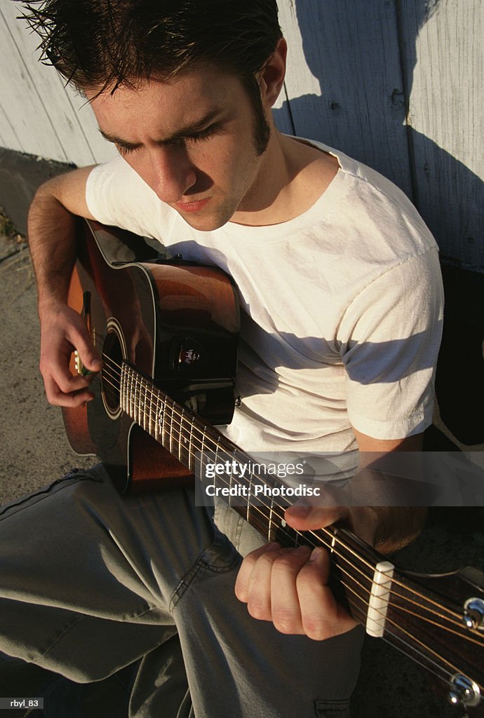 A young man in a white t-shirt and tan pants is sitting on a sidewalk playing his guitar