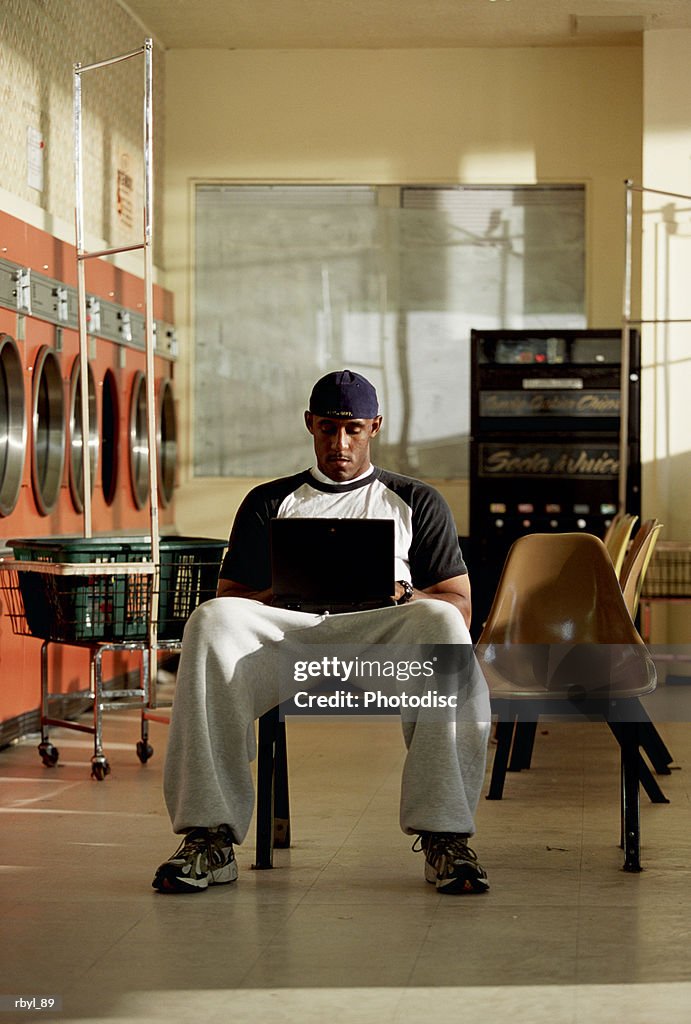 A young man in grey sweatpants and a t-shirt is sitting in a laundromat typing on a laptop computer