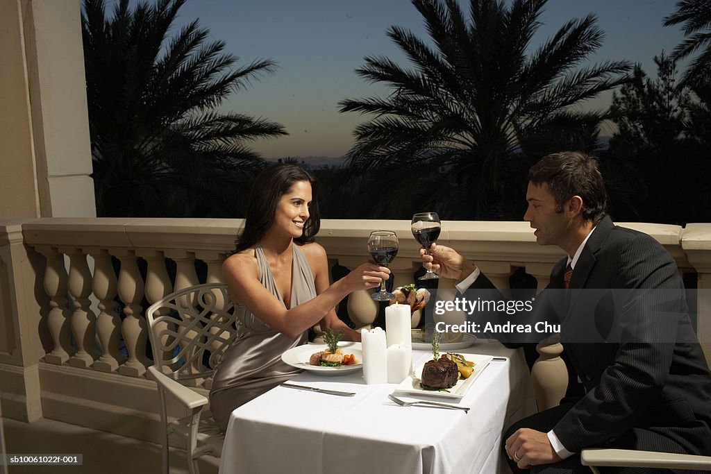 Couple in evening wear having dinner on balcony, toasting with red wine