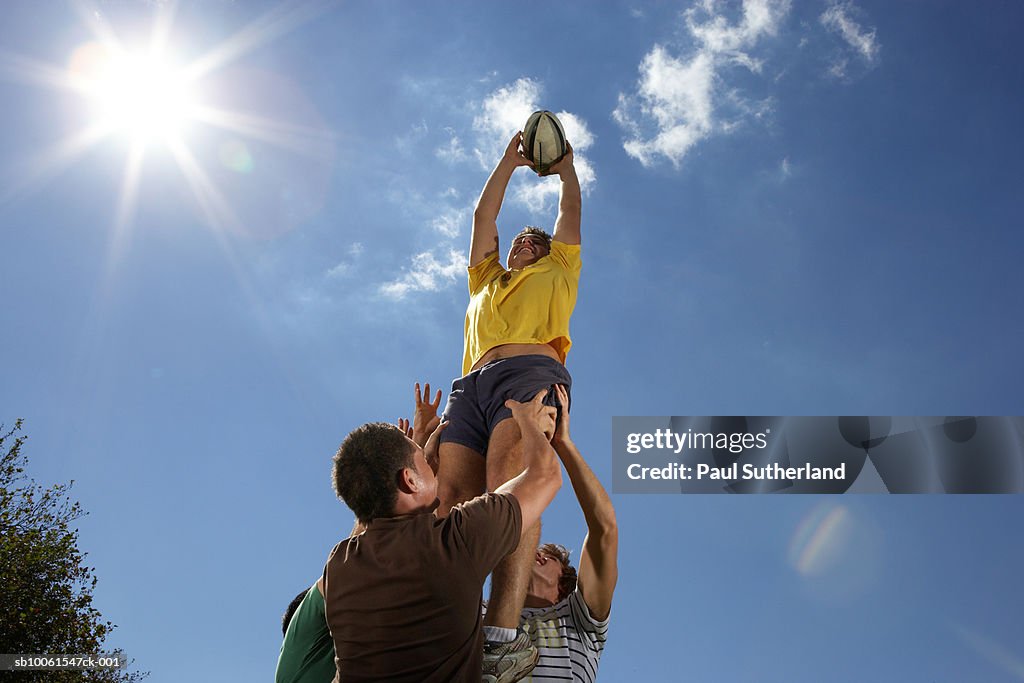 Men playing rugby, holding man with ball up in air, low angle view