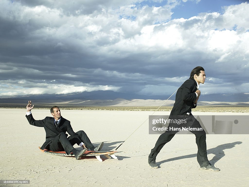 Businessman pulling another businessman sitting on sled in desert