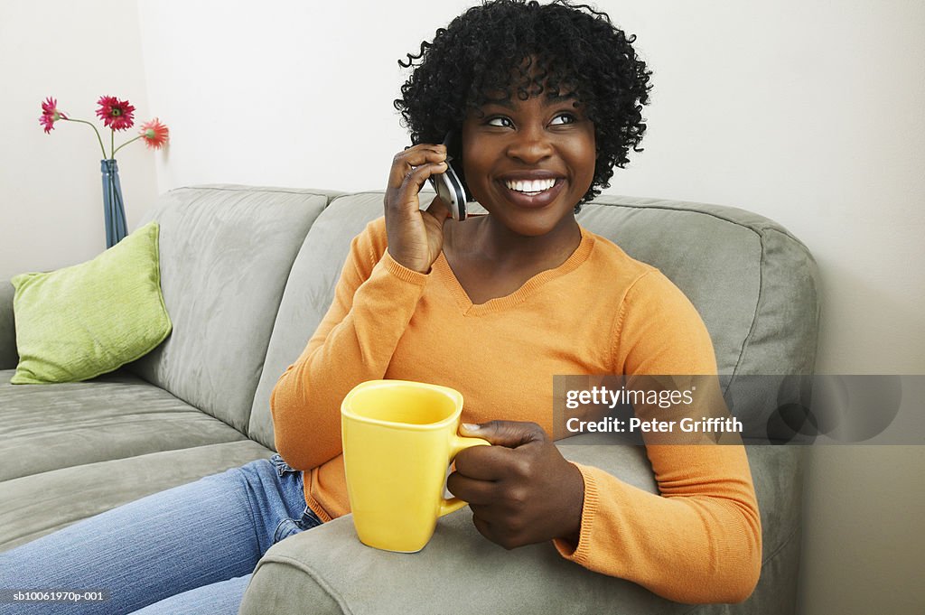 Young woman sitting on sofa, holding mug and using mobile phone