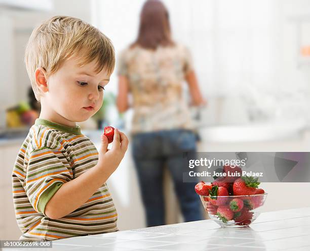 boy (3-4) eating strawberries, while mother cooking food in background - child eating a fruit stockfoto's en -beelden