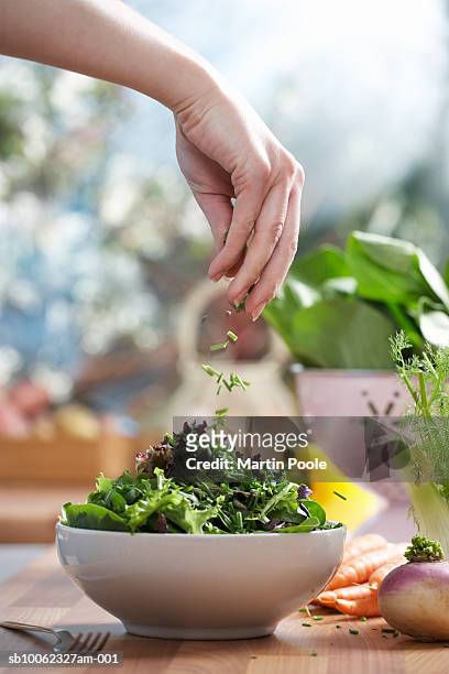 woman pouring herbs into bowl of salad in kitchen, close-up of hand - saladekom stockfoto's en -beelden
