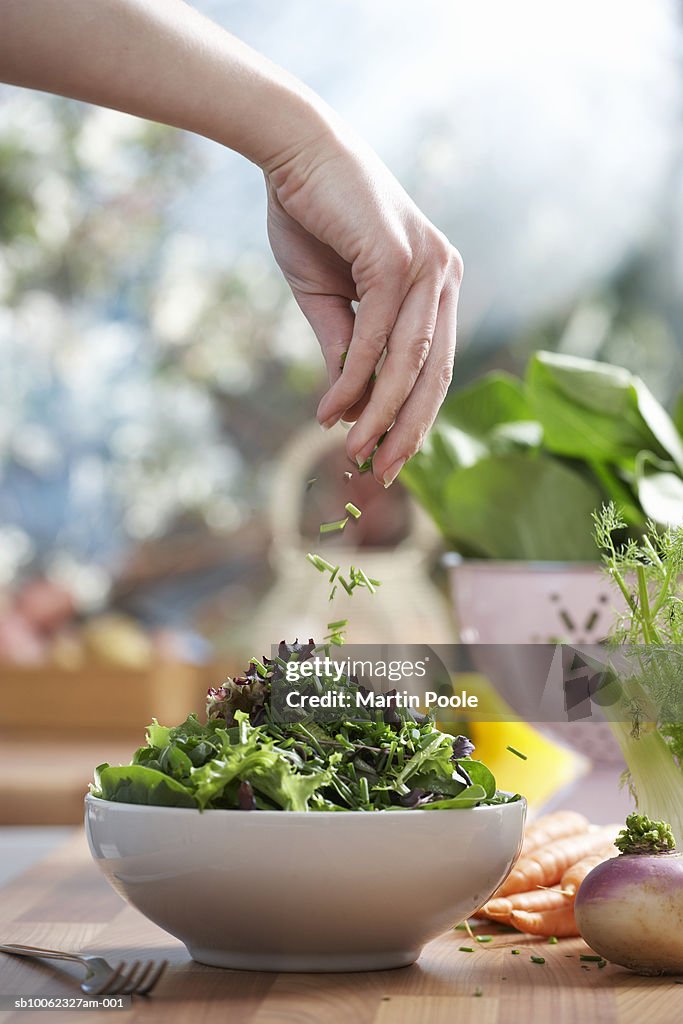 Woman pouring herbs into bowl of salad in kitchen, close-up of hand