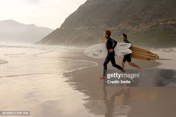 father and son (14-15) carrying surfboard, running on beach - barefoot teen boys stock pictures, royalty-free photos & images