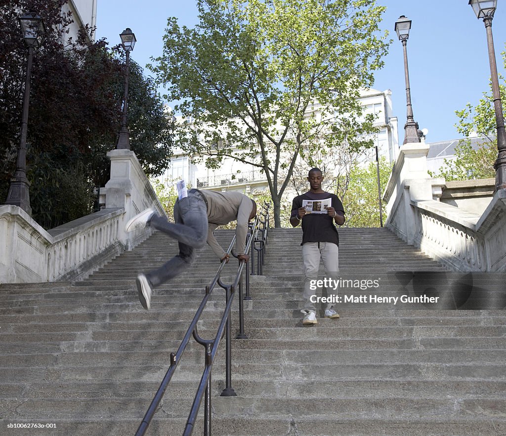 Man walking down steps reading newspaper, another man jumping over railings