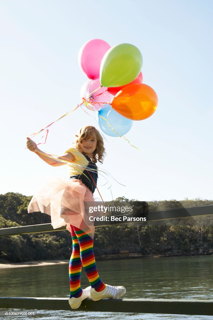 Girl (8-9) standing on fence by river, holding balloons, portrait